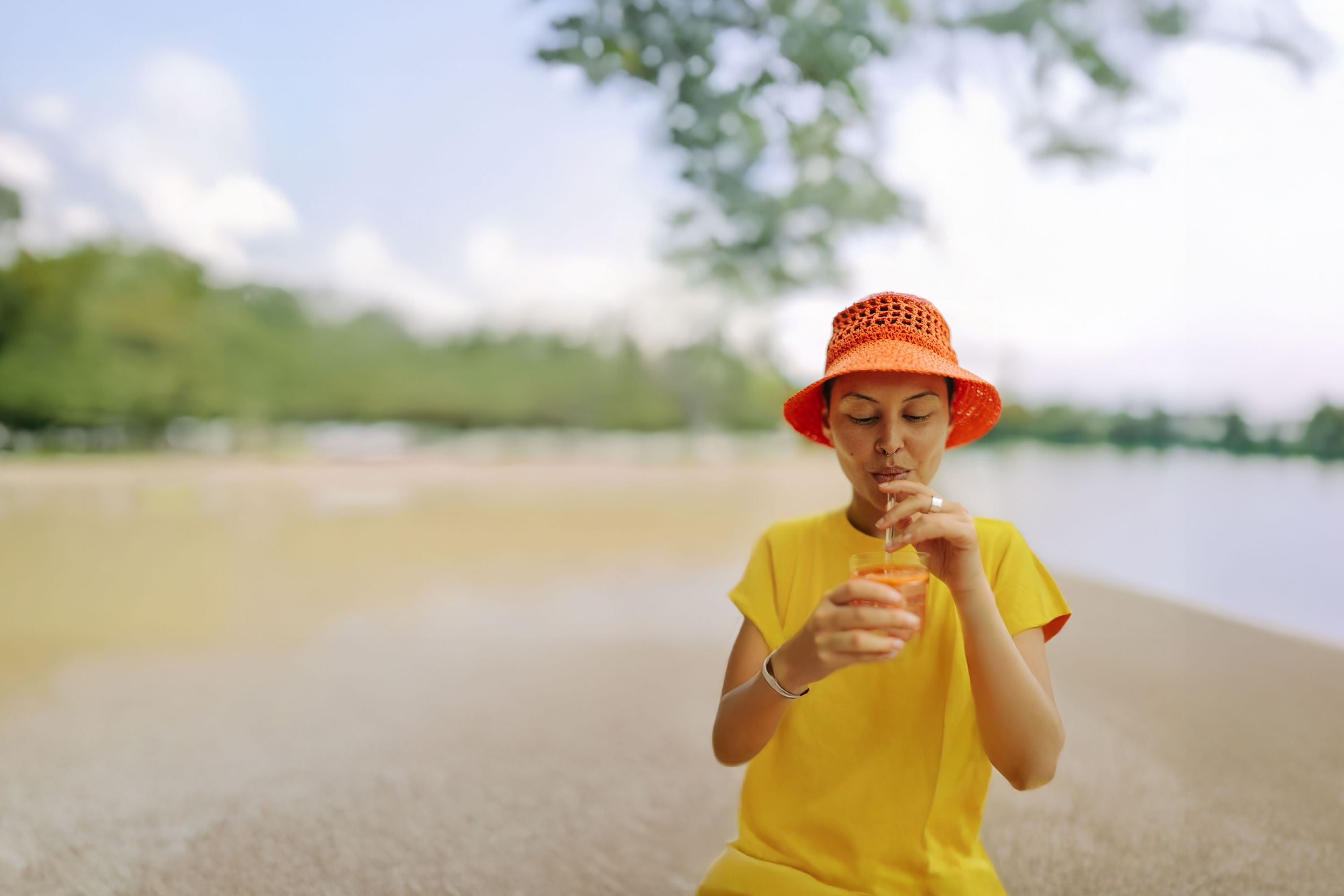 „Frau mit gelbem Shirt und orangem Hut steht am Sandstrand und hält ein Glas mit Eistee in der Hand, im Hintergrund unscharfes Grün und Wasser