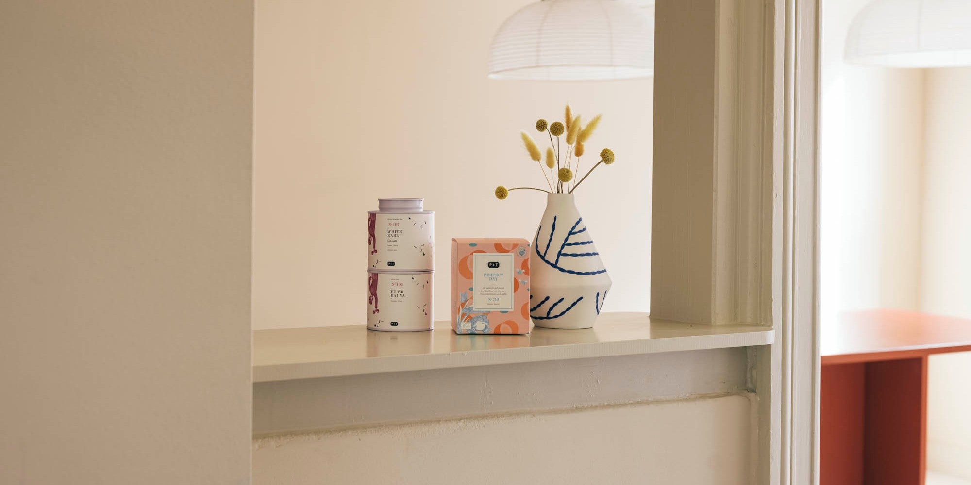 Zwei gestapelte weiße Teedosen, eine orangefarbene quadratische Teeverpackung und eine Vase mit gelben Kugelblumen vor beiger Wand