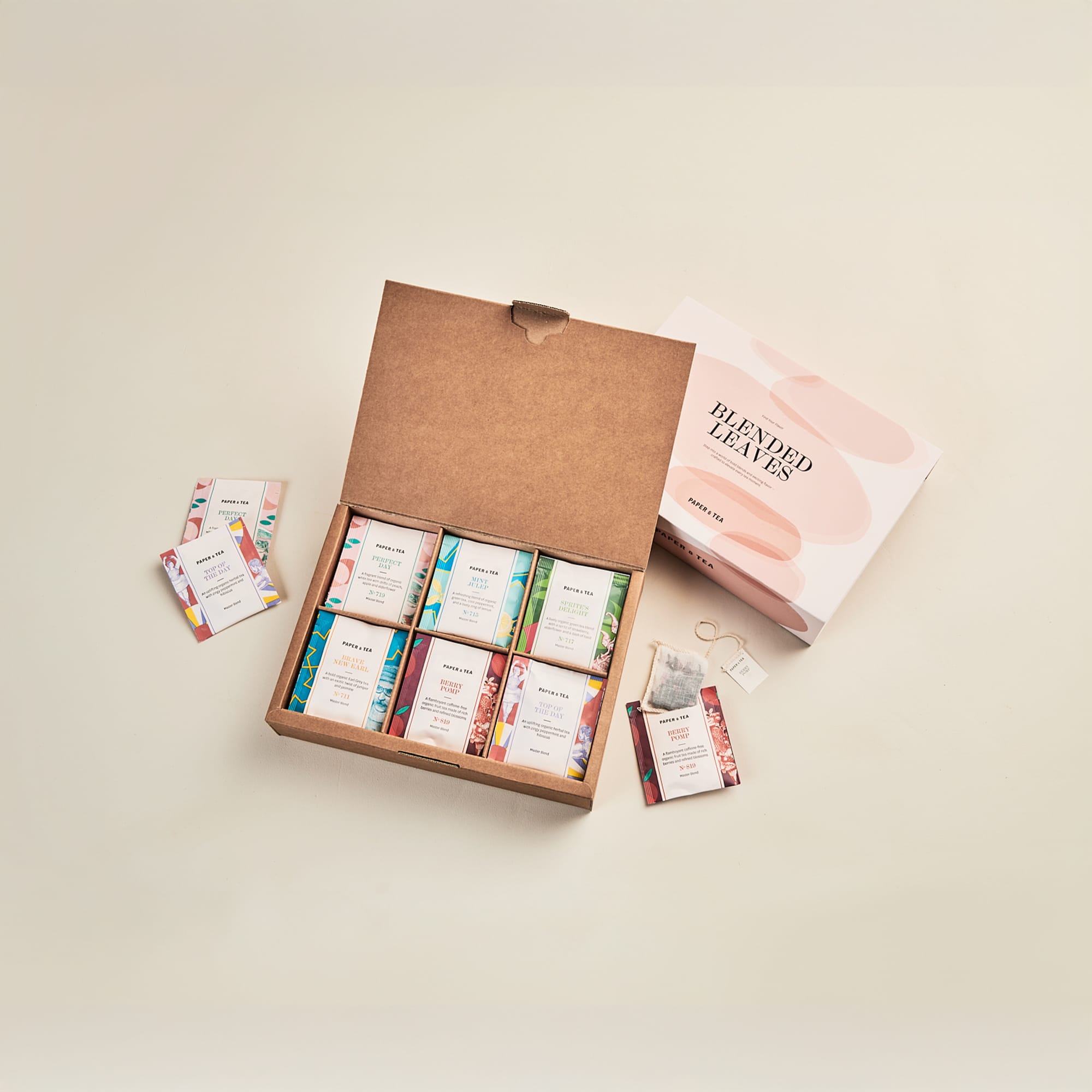 Geöffnete Papier & Tee Schachtel mit acht bunten Teepackungen und zwei Teebeuteln auf hellem Hintergrund.