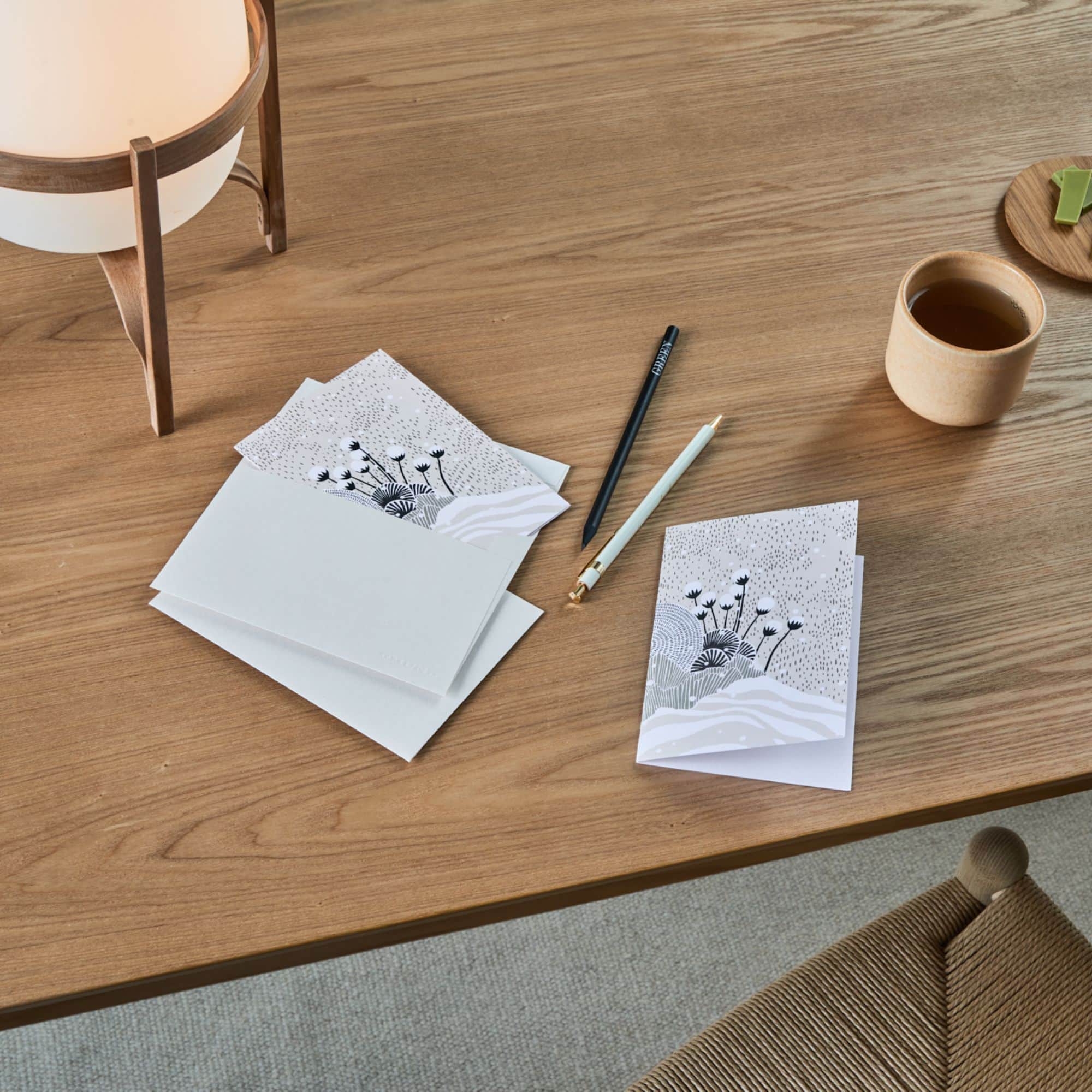 Holztisch mit floralen Grußkarten, beigem Teebecher, Stiften und grünem Snack.
