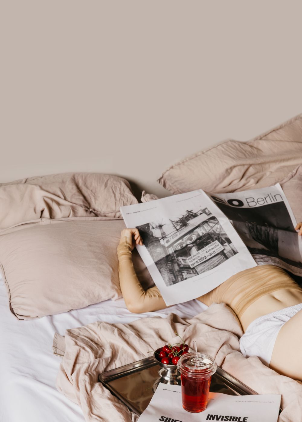 Person liest auf einem Bett mit beiger Bettwäsche eine Zeitung, frische Kirschen und rotes Getränk daneben