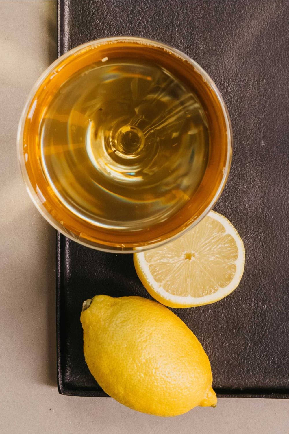 Obenaufnahme eines Glases mit goldenem Tee, daneben ganze und halbierte Zitrone auf dunklem Tablett.