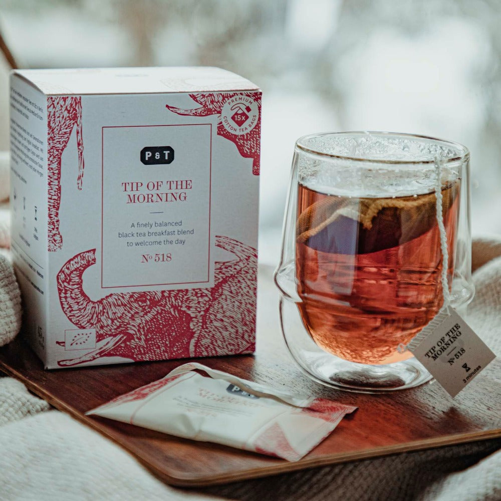 Holztisch mit weiß-rotem Teekarton, Glastasse mit 'TIP OF THE MORNING' Tee und einzeln verpacktem Teebeutel.