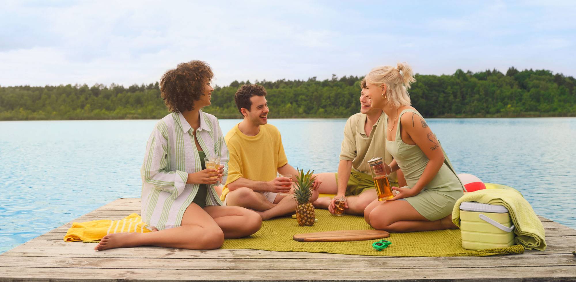 Vier junge Erwachsene auf einem Steg am See mit Ananas und Getränken auf einer Picknickmatte.
