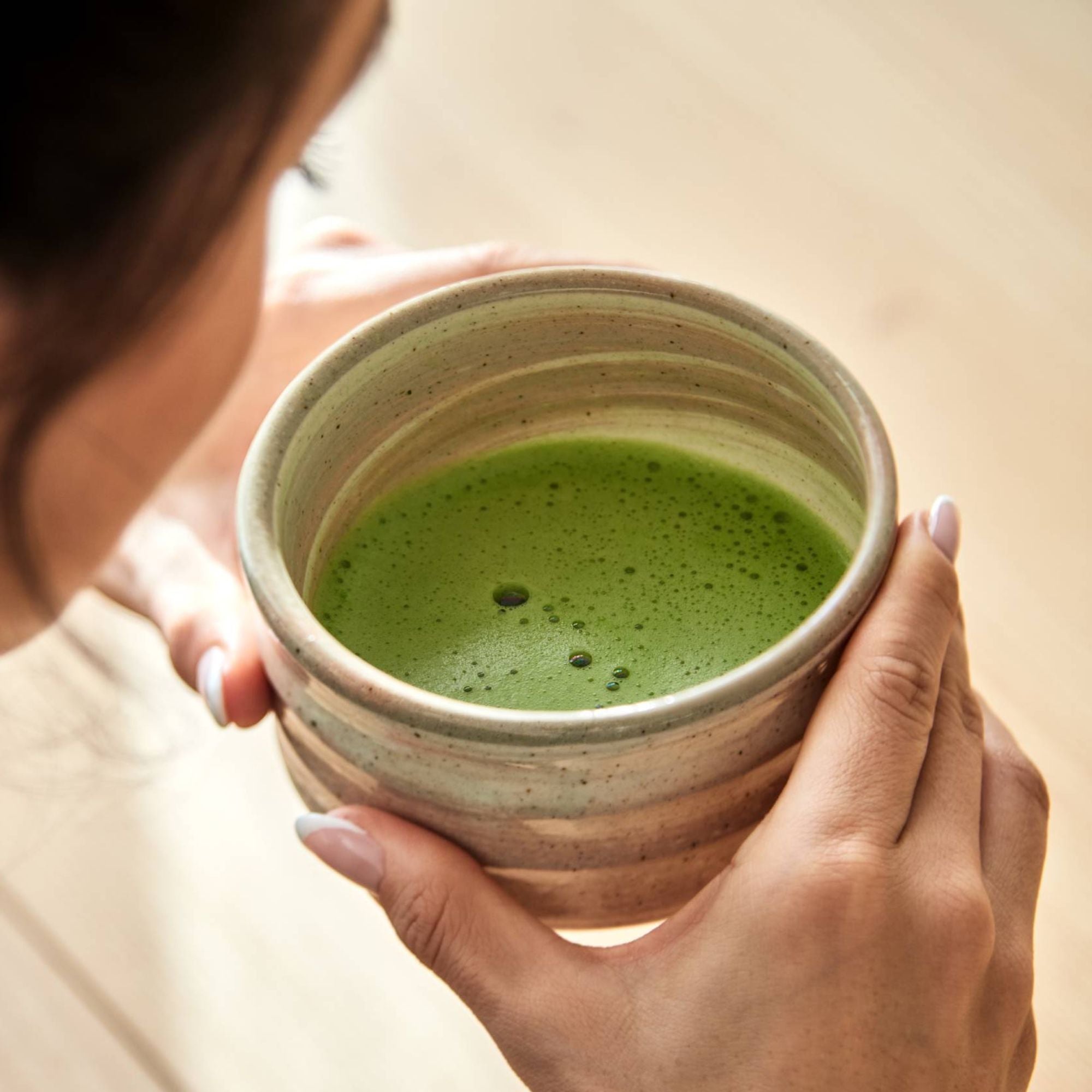 Hände halten einen rustikalen Keramikbecher mit schaumigem, lebendig grünem Matcha-Tee vor hellem Holz.