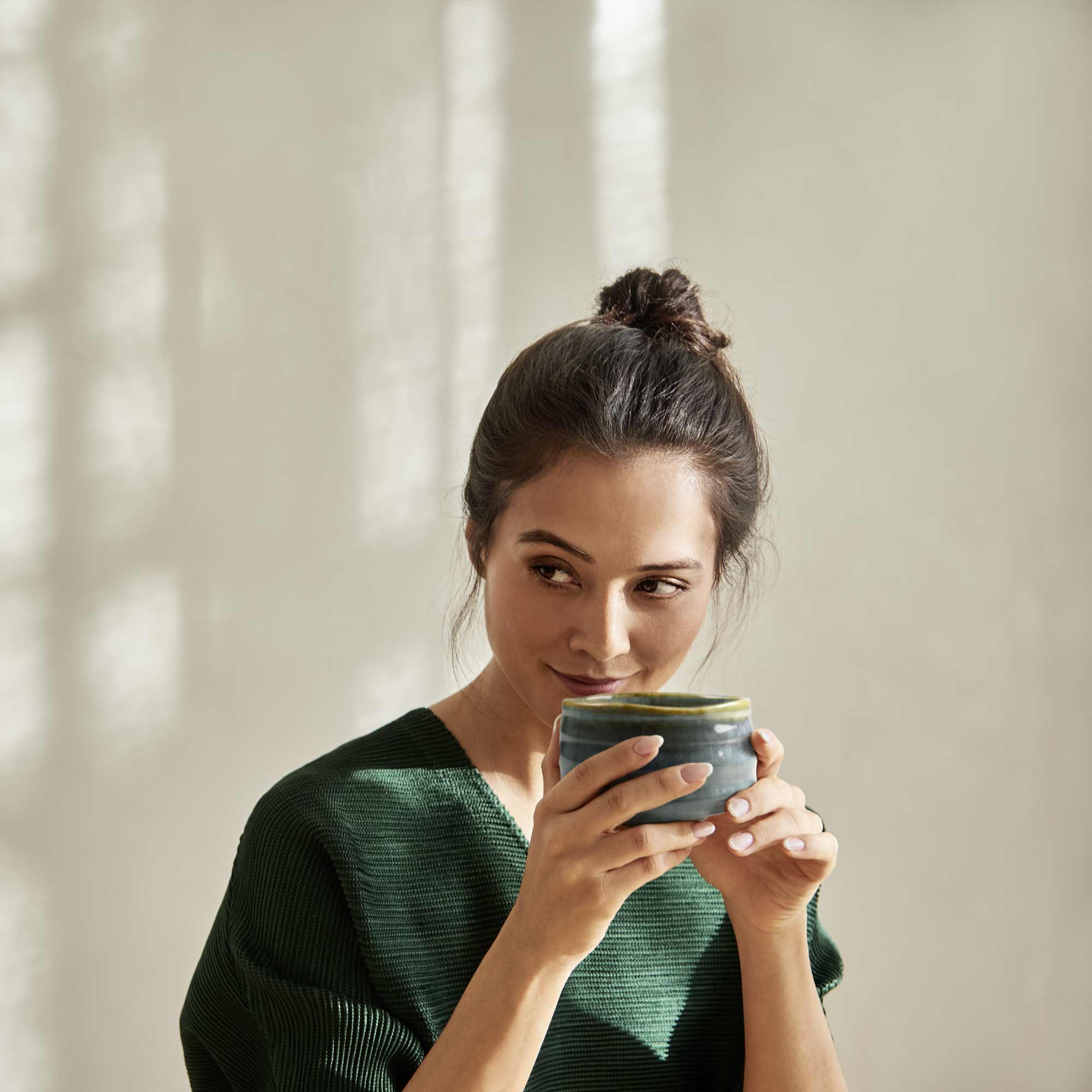 Junge Frau mit dunklem Dutt und grünem Oberteil hält lächelnd eine blaue Keramiktasse vor beige beleuchteter Wand.