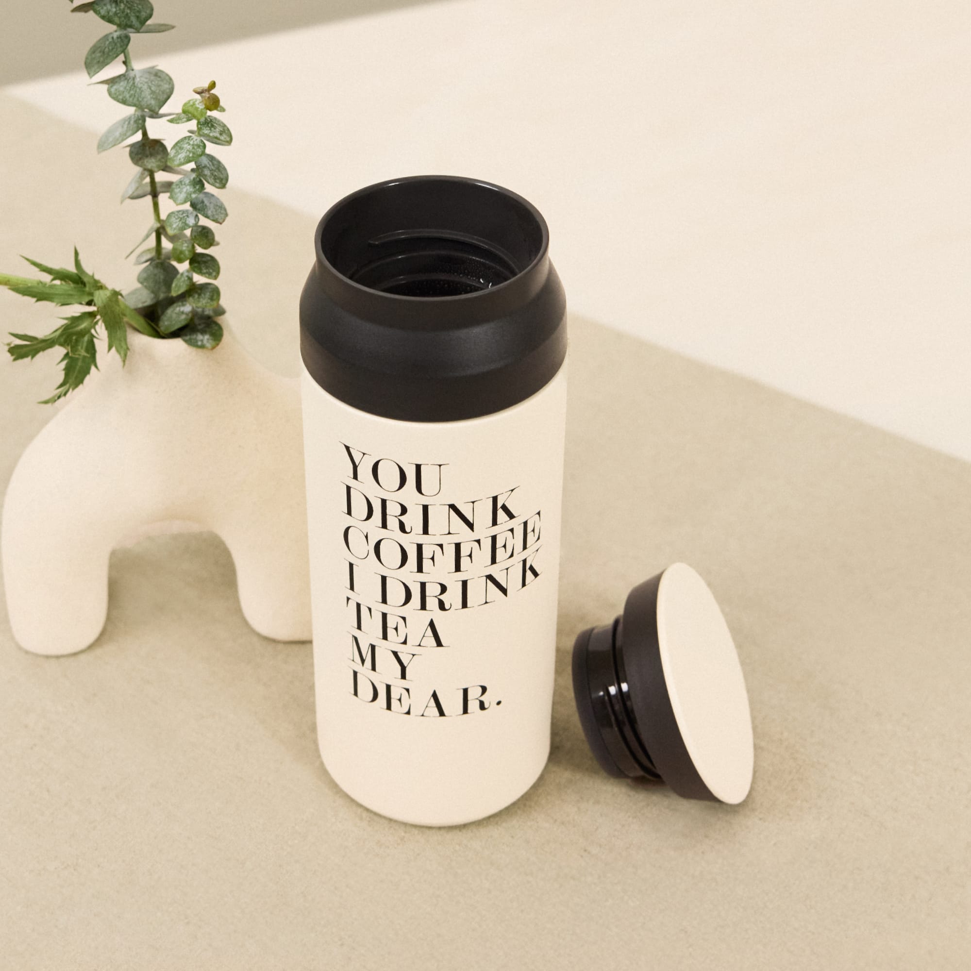 Eleganter weiß-schwarzer Isolierbecher mit Kaffee-Spruch auf Tisch neben Vase mit Eukalyptuszweigen.