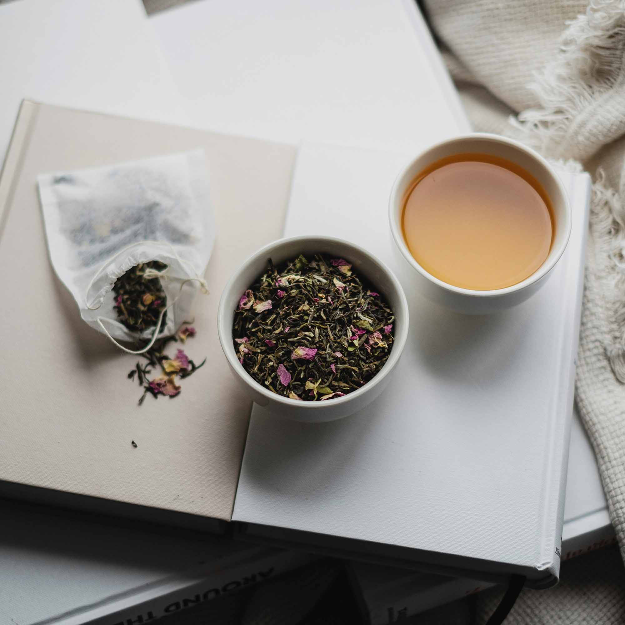 Weiße Keramiktasse mit hellem Tee neben Schale mit Teeblättern und Blüten auf gestapelten Büchern.