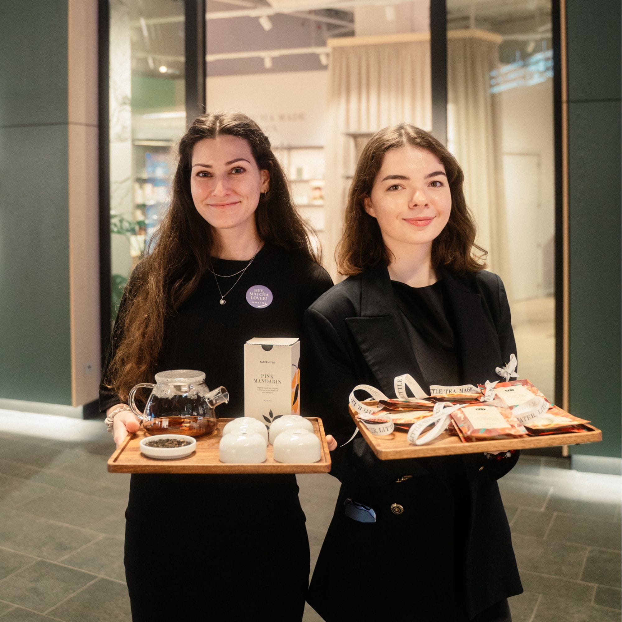 Zwei Frauen zeigen Tabletts mit Paper & Tea Pink Mandarin und Little Tea Magic in einem modernen Teeladen.