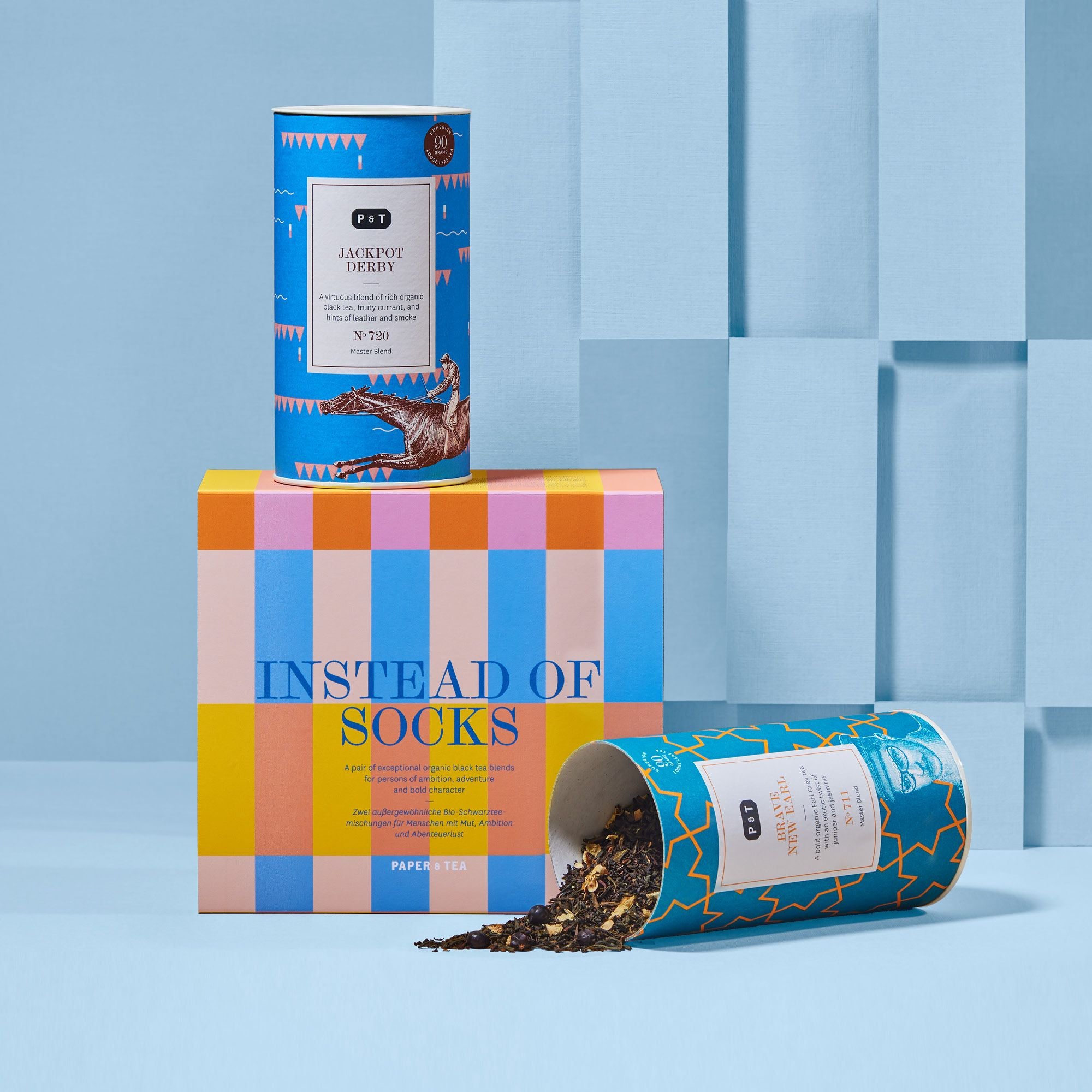 Zwei Bio-Schwarztee-Dosen von Paper & Tea mit Pferde-Design und Muster, neben bunter Geschenkbox vor blauem Hintergrund.