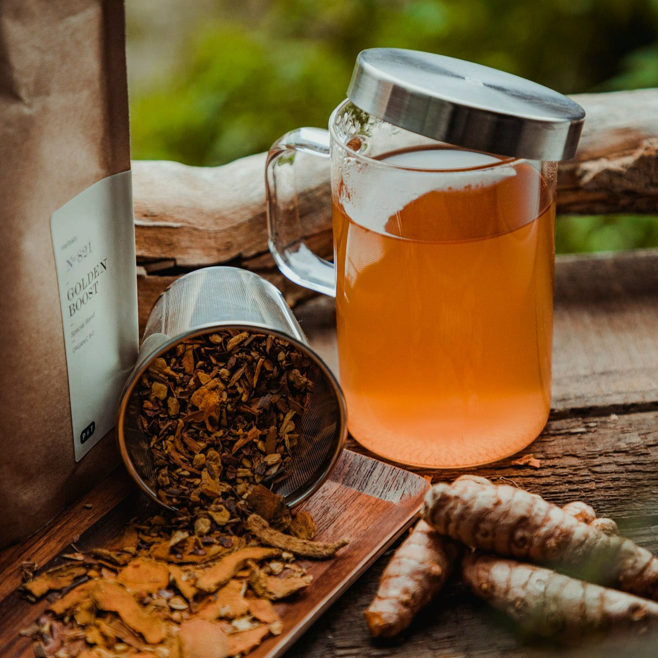 Klarer Glasmug mit GOLDEN BOOST Tee, metallischem Deckel, getrockneten Kräutern und frischen Kurkumawurzeln.