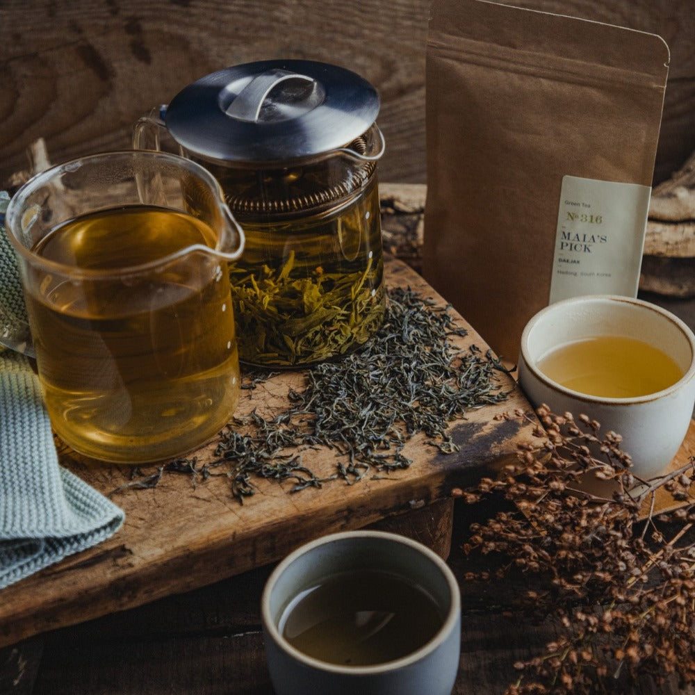 Rustikales Teeset mit Glasteekanne, grünem Tee, Keramikbecher und brauner Verpackung auf Holzbrett.