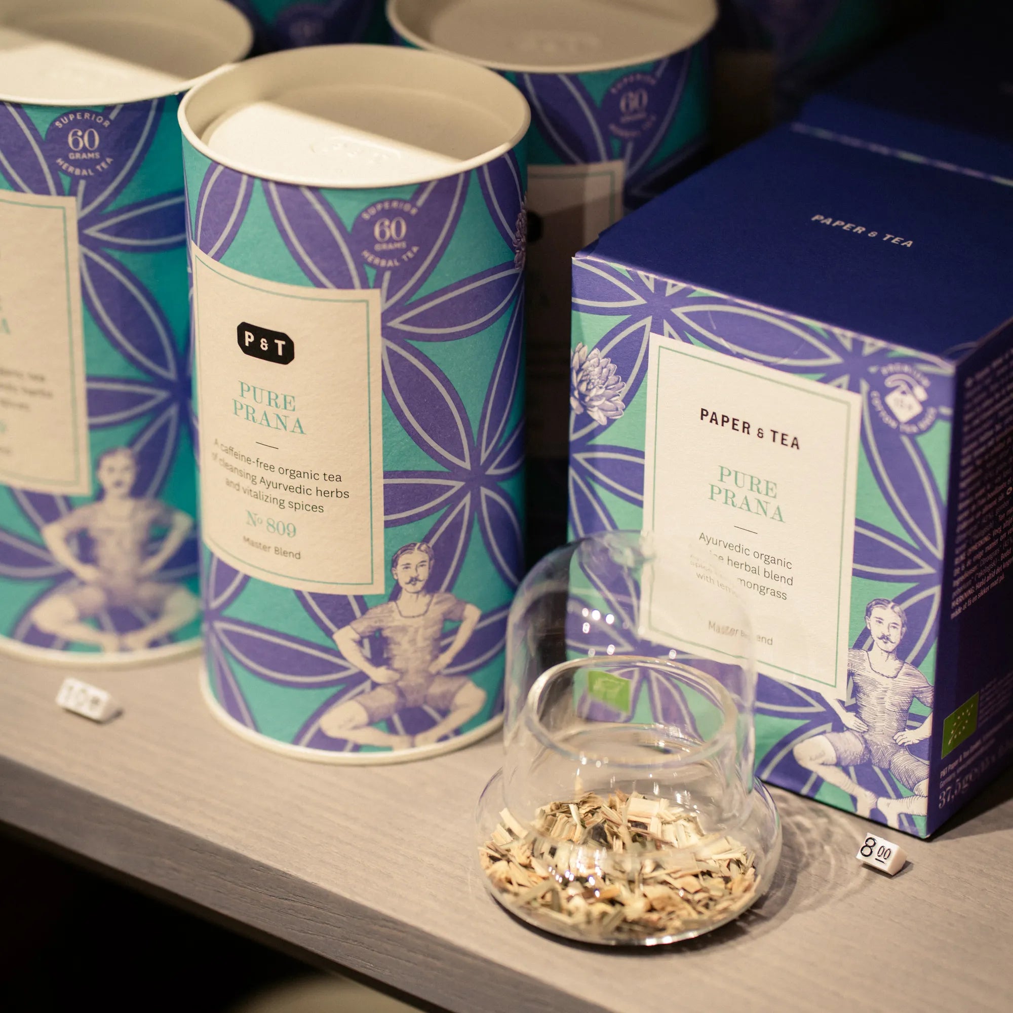 PURE PRANA Kräutertee von Paper & Tea in hochwertiger Verpackung neben Glas mit Teekräutern.