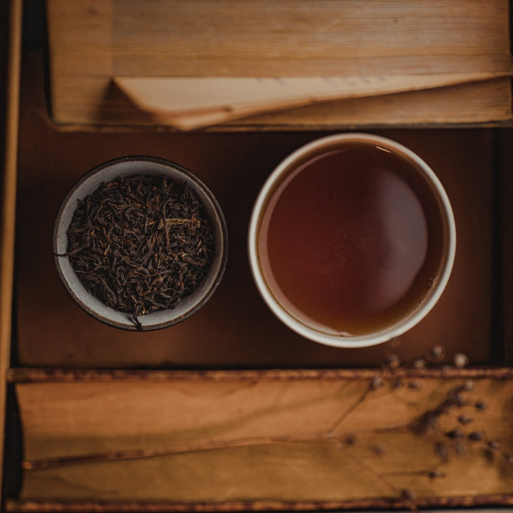 Keramikschale mit losen Teeblättern und Tasse bernsteinfarbenen Tees auf Holztablett mit Papierseiten.