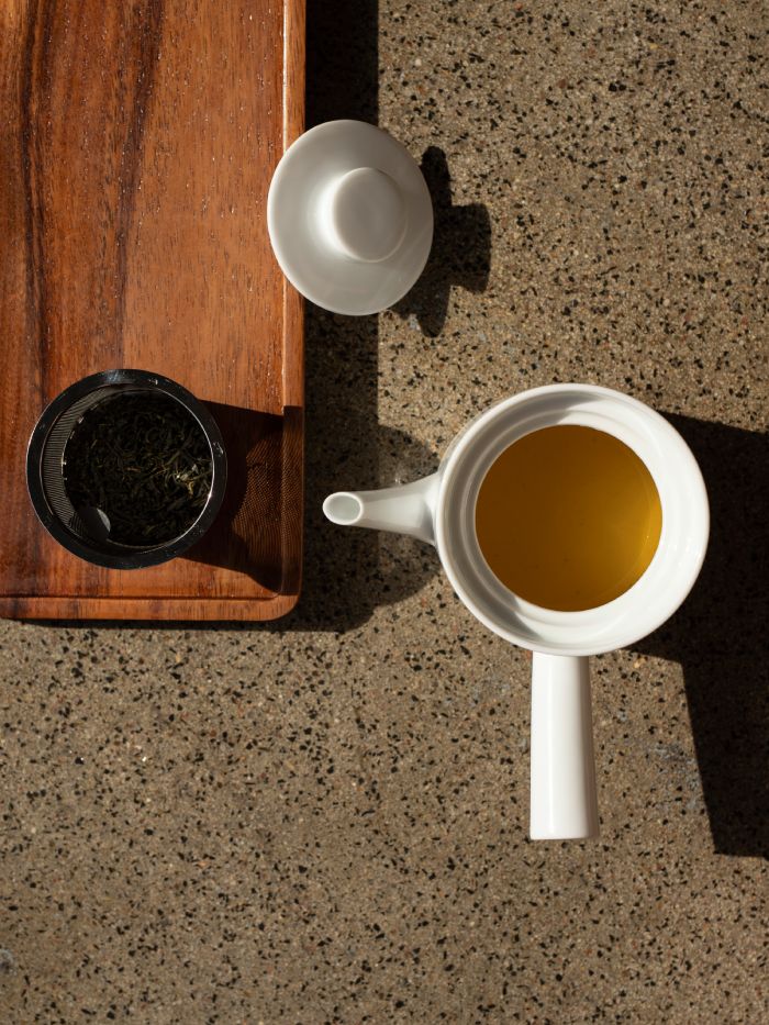 Weißer Keramik-Teekessel mit gelbem Tee und schwarzer Teedose auf Holztablett.