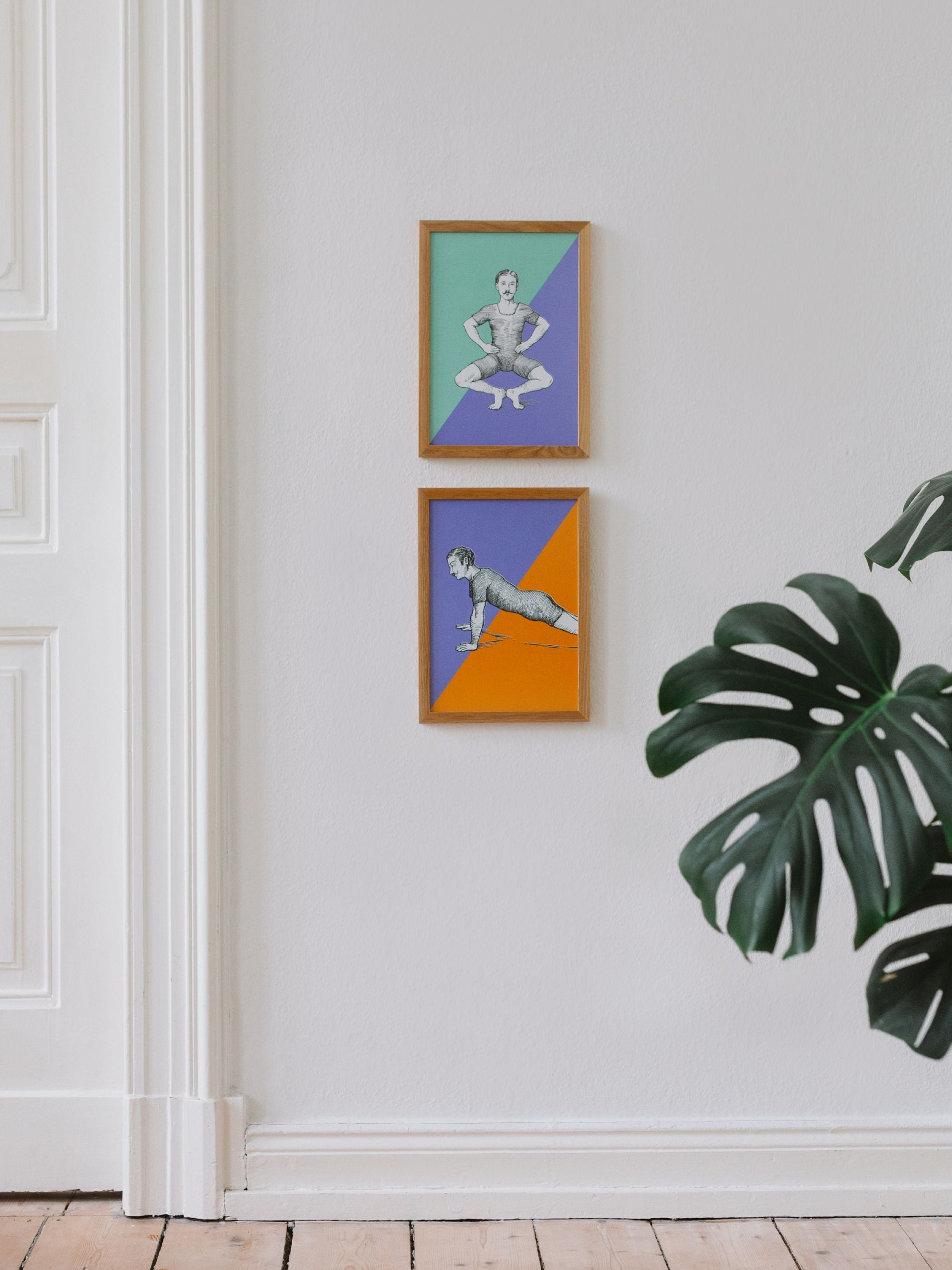Zwei Holzrahmen mit Vintage-Übungsillustrationen vor geometrischen Hintergründen, neben weißer Tür und Monstera-Pflanze.