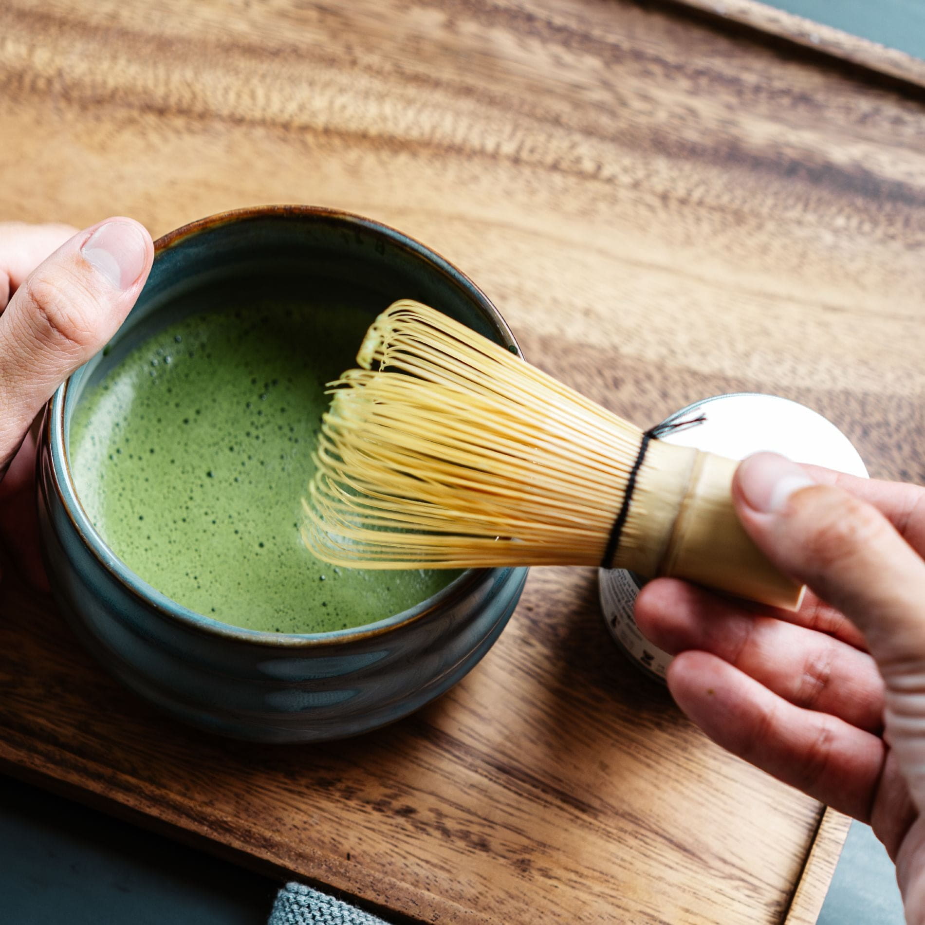 Hand rührt cremigen grünen Matcha-Tee in Keramikschale mit Bambusbesen auf Holztablett.