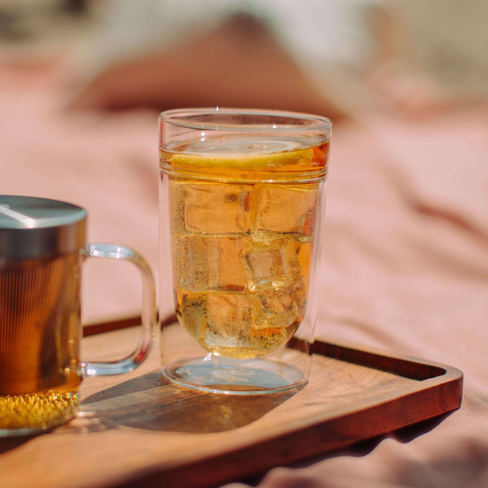 Ein erfrischender Eistee mit Zitronenscheibe in einem doppelwandigen Glas auf einem Holztablett mit Teesieb vor rosa Hintergrund.