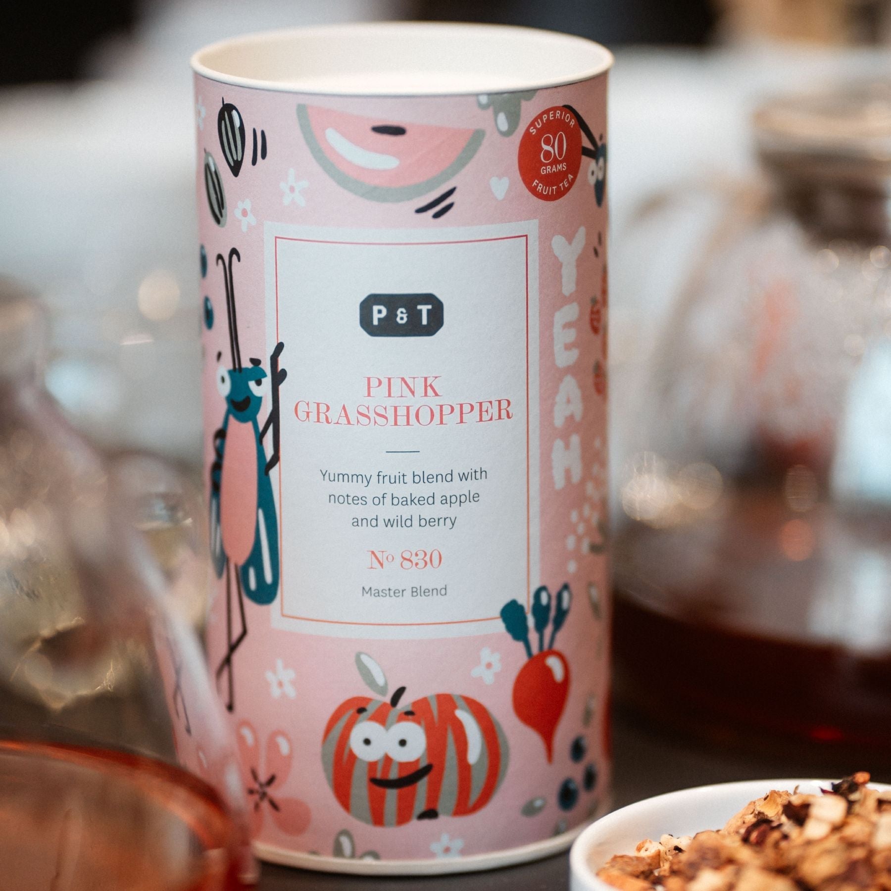 Pinke Teedose von P & T mit Illustrationen eines blauen Grashüpfers und Früchten, Tee mit Apfel- und Beerennoten.