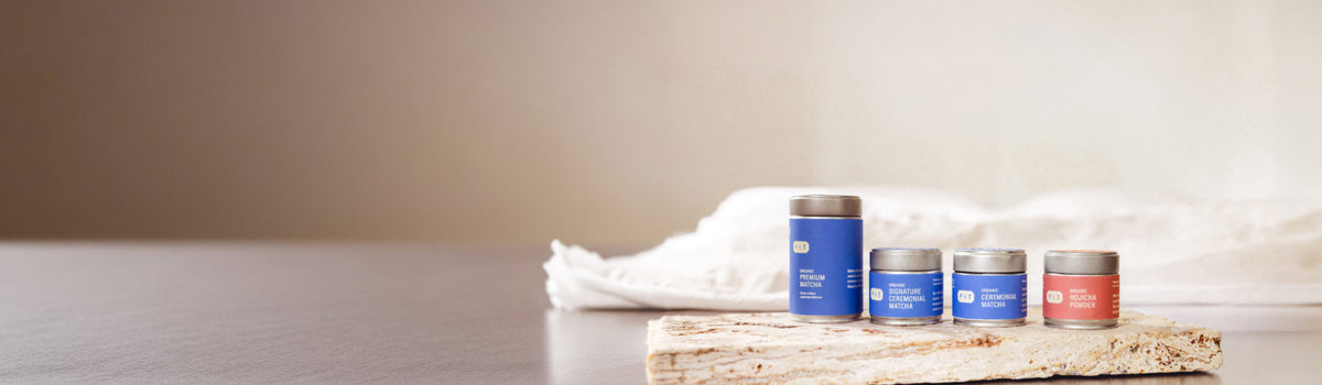 Vier Dosen mit Bio-Teepulver von P&T in Pastellblau und Rosa auf Naturstein, einschließlich Matcha und Hojicha.