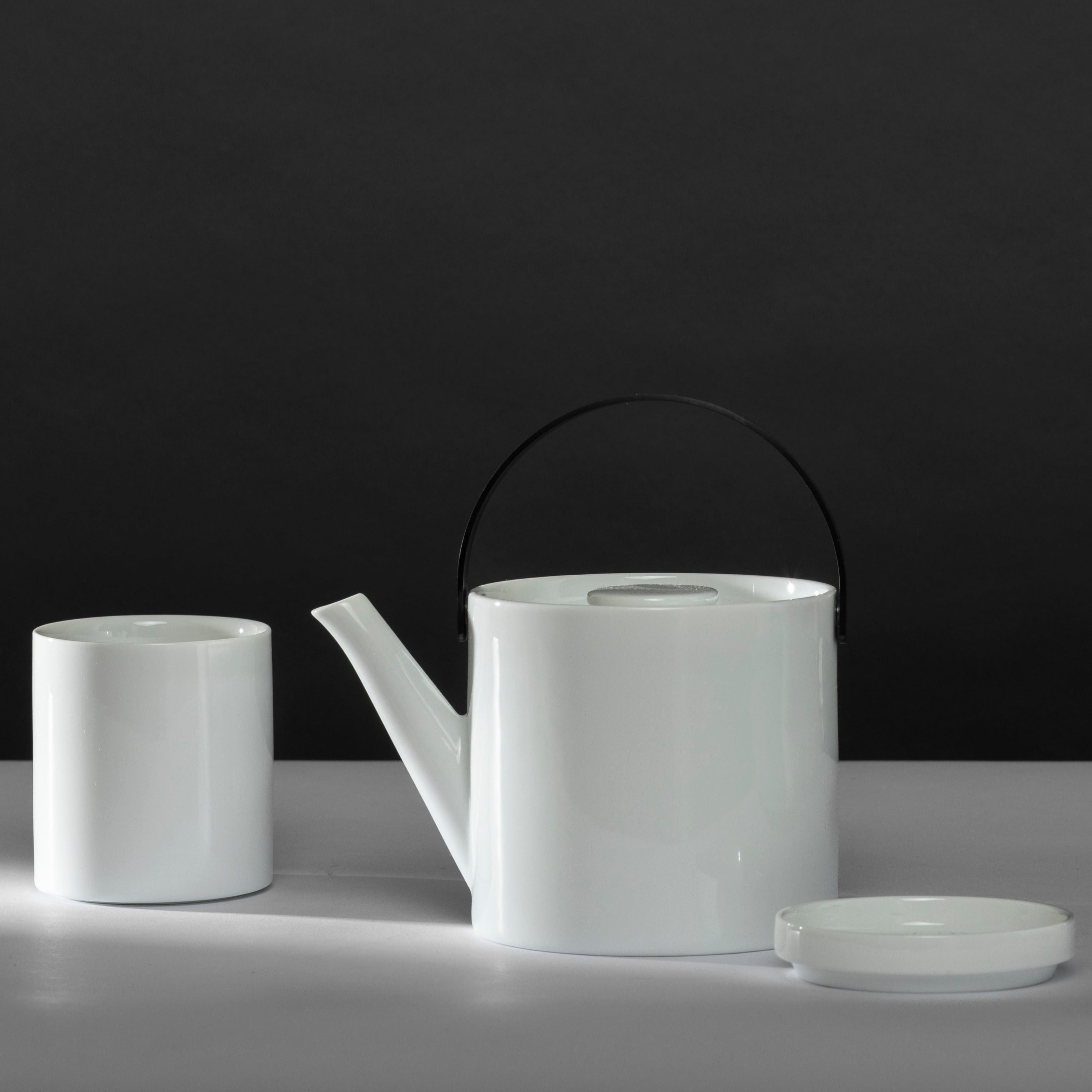 Weißer Keramik-Teekessel mit schwarzem Griff, daneben weiße zylindrische Tasse und passende Untertasse vor grauem Hintergrund.
