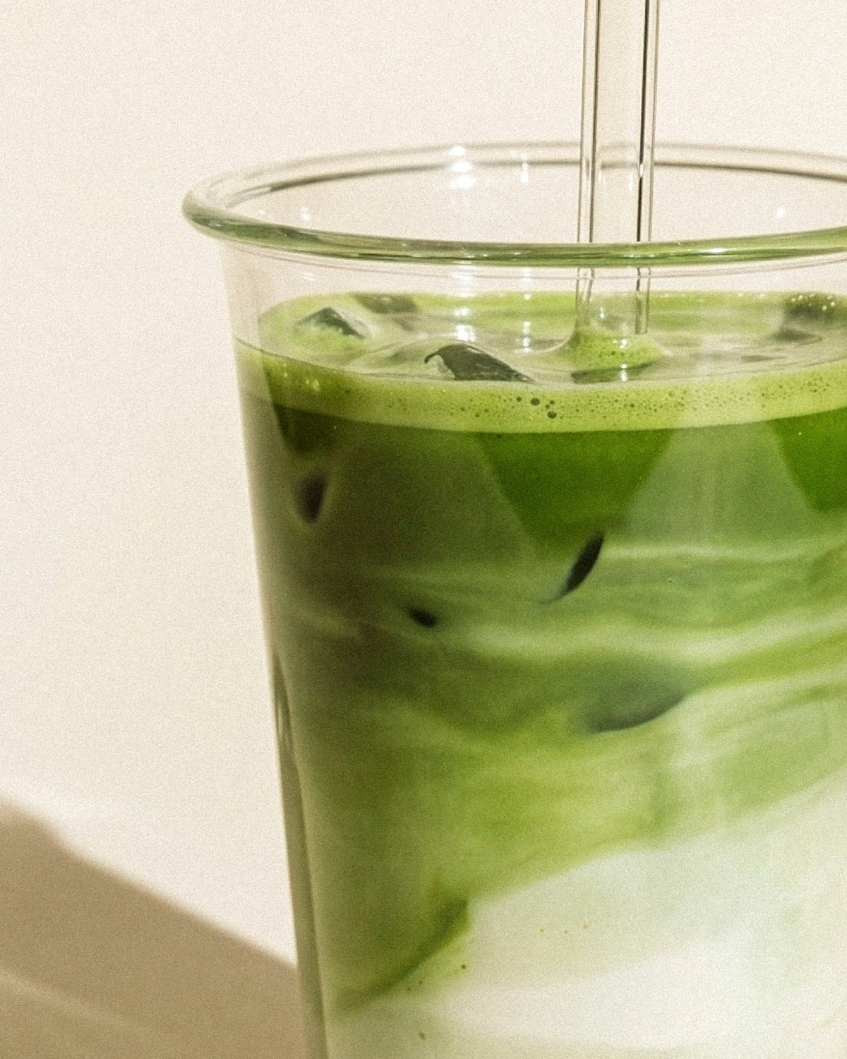 Nahaufnahme eines durchsichtigen Bechers mit grünem Matcha-Eistee, Milch, Eiswürfeln und zwei Strohhalmen.