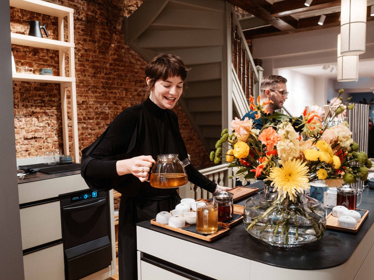 Junge Frau gießt Tee aus einer Glasteekanne in kleinen Becher auf einem Holztablett in gemütlicher Küche.