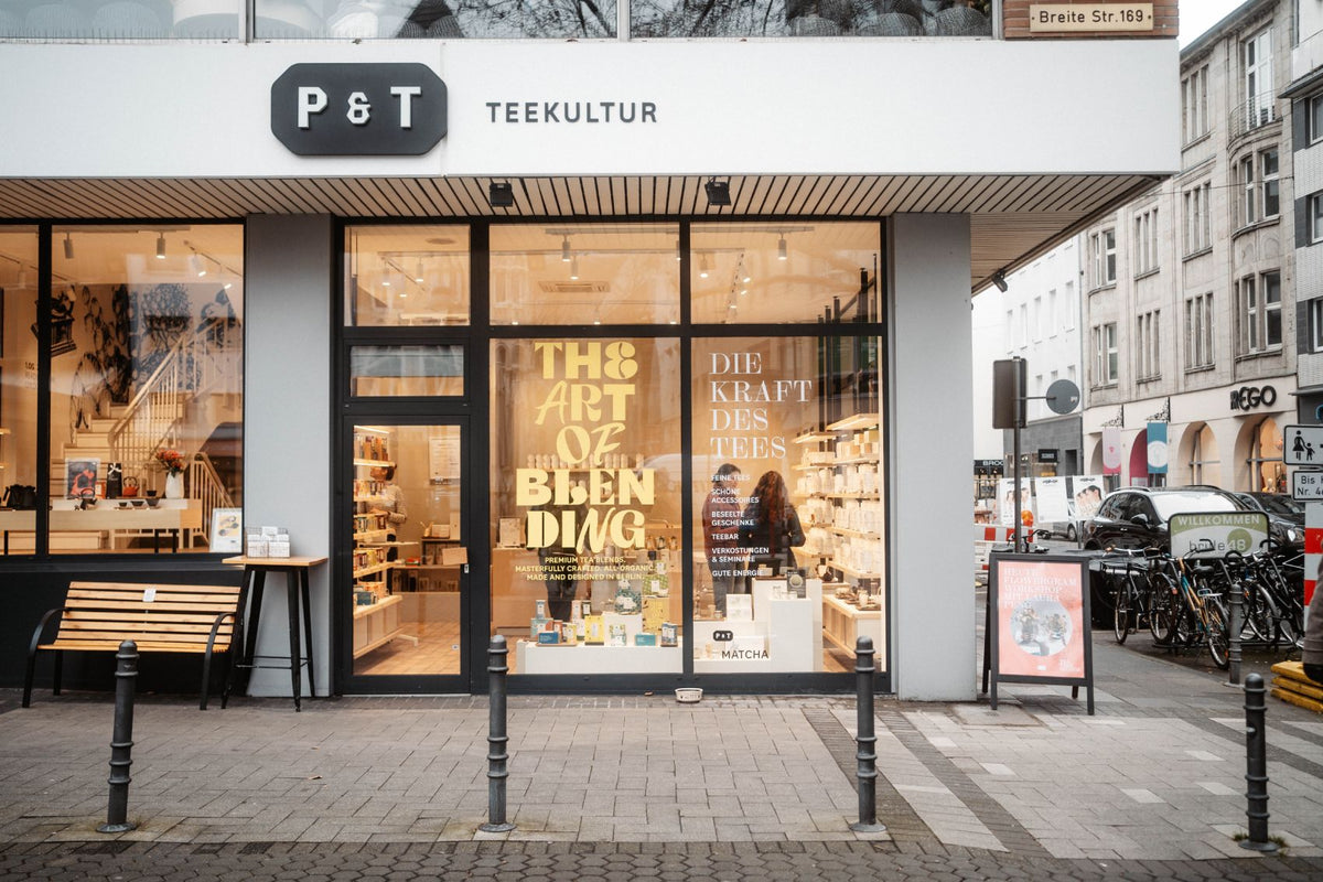 P & T TEEKULTUR Laden mit großen Glasfenstern, Holzbänken und Straßenschild Breite Str. 169.