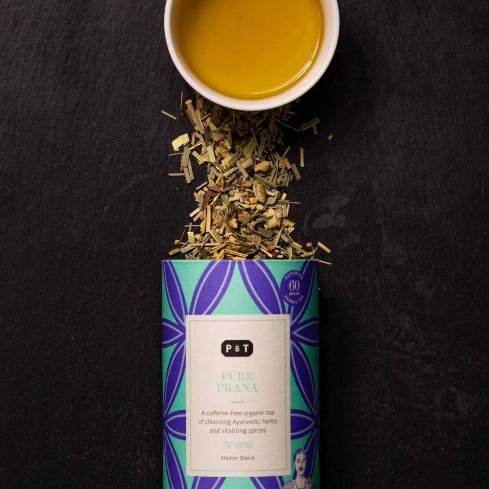 Geöffnete zylindrische Dose PURE PRANA Kräutertee mit ayurvedischen Kräutern und Tasse goldgelbem Tee.
