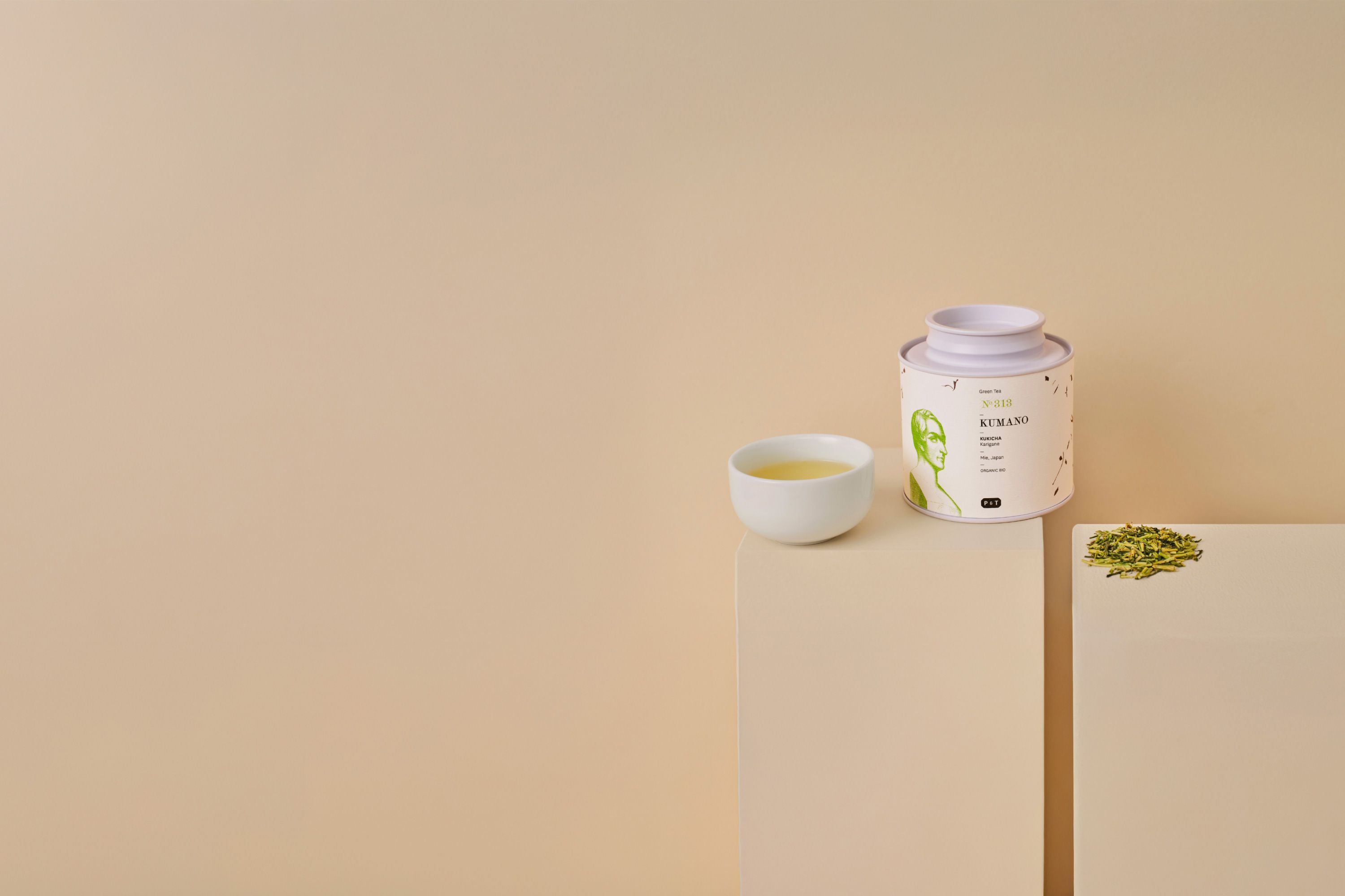 Weiße Dose Kumano Kukicha Karigane Tee und Tasse mit grünem Tee vor beigem Hintergrund.