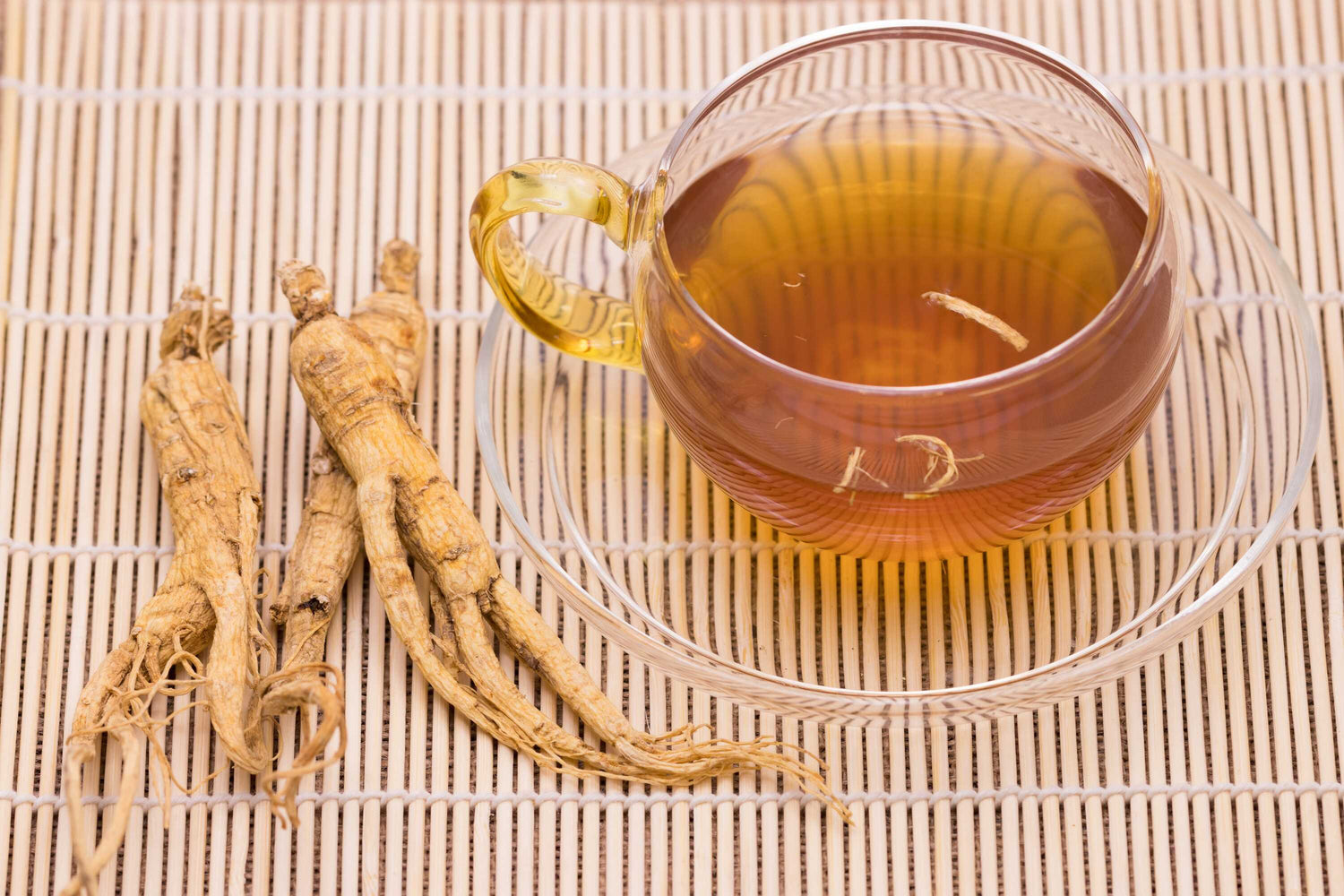 Ein Glas mit braunem Ginseng-Kräutertee auf einer Bambusmatte neben getrockneten Ginsengwurzeln