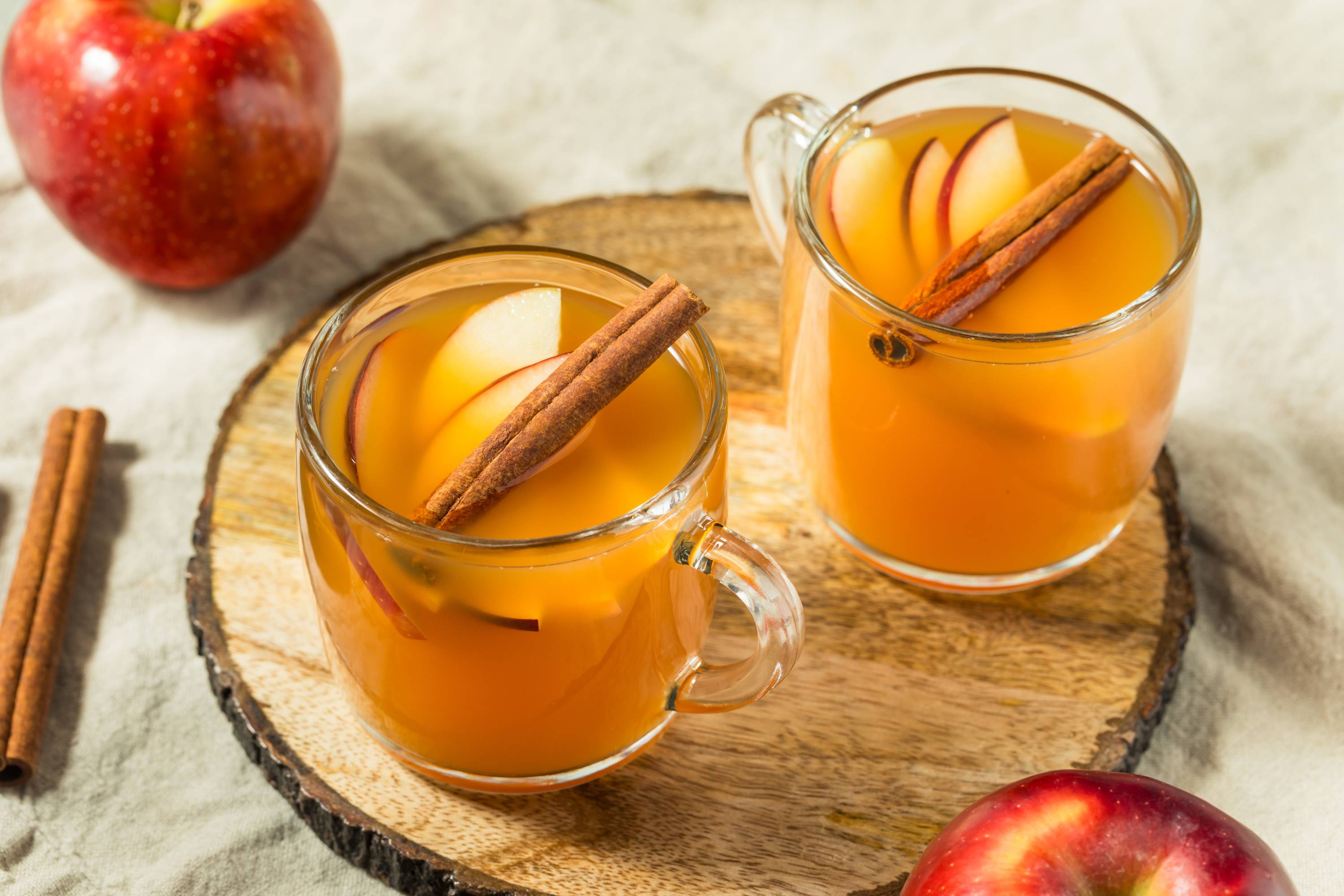 Zwei transparente Gläser mit warmem Winterpunsch mit Apfelsaft, Orangensaft und Zimtstangen auf rundem Holzbrett