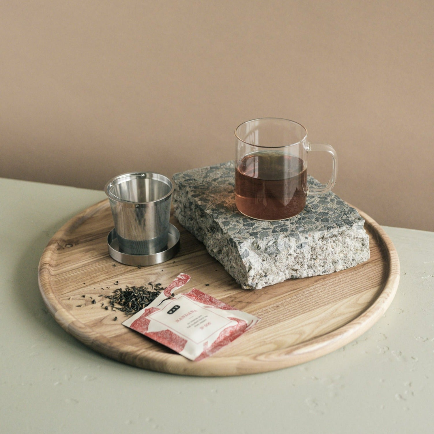 Rundes Holztablett mit Glasbecher dunklen Tee, Steinblock, Edelstahl-Teesieb und rotem Beutel losen Blättern.