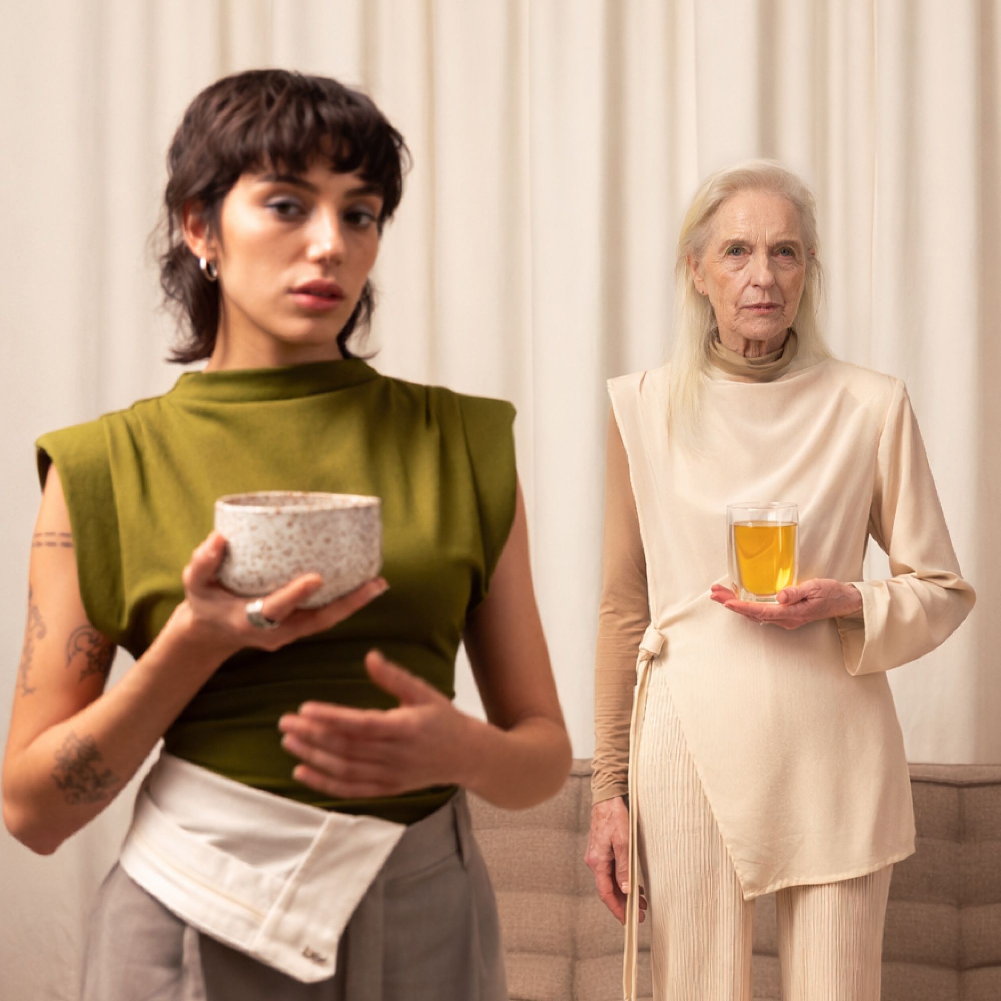Zwei Frauen in neutraler Umgebung: Jüngere mit Tattoos hält Keramikbecher, Ältere mit Glasbecher und beigem Outfit.