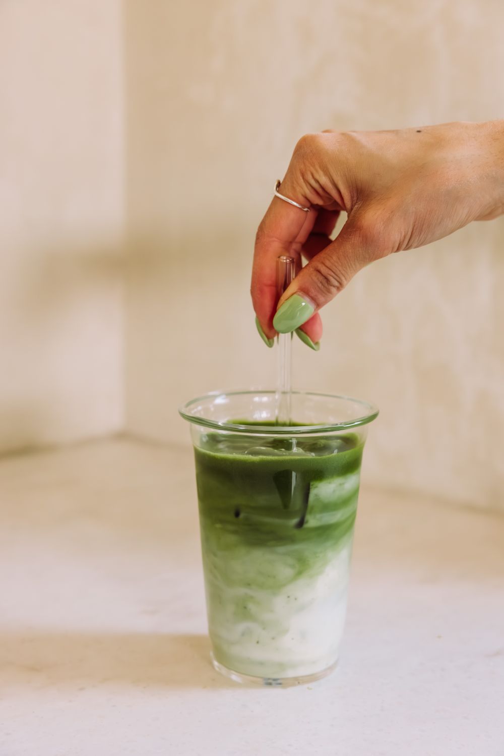 Nahaufnahme eines Eistee-Matcha-Latte mit weißen Milch- und grünen Matcha-Schichten, gerührt von einer Hand mit hellgrünem Nagellack.