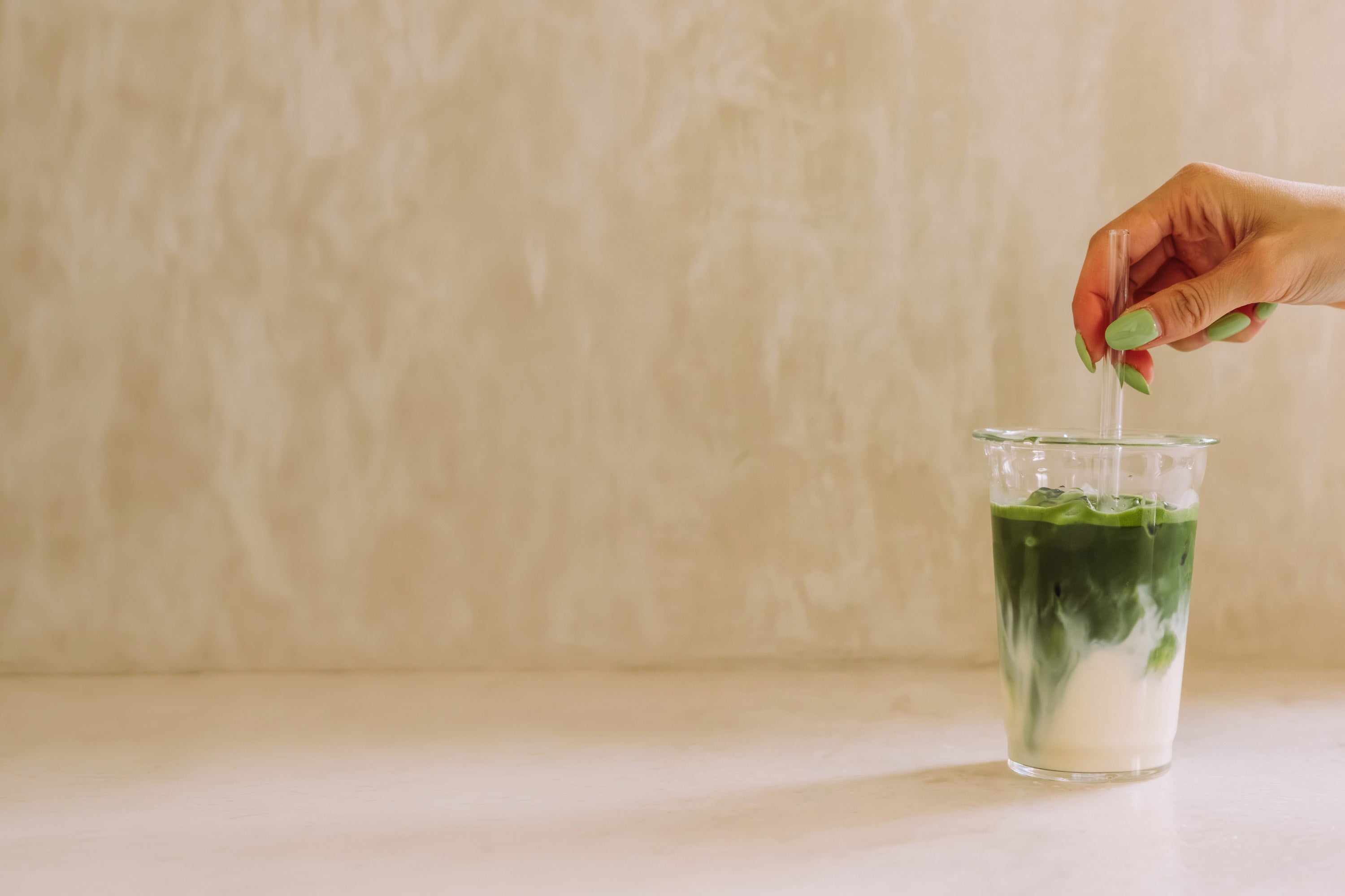 Glas Eistee mit weißer Milch und grünem Matcha, gerührt mit Strohhalm vor beigem Hintergrund.