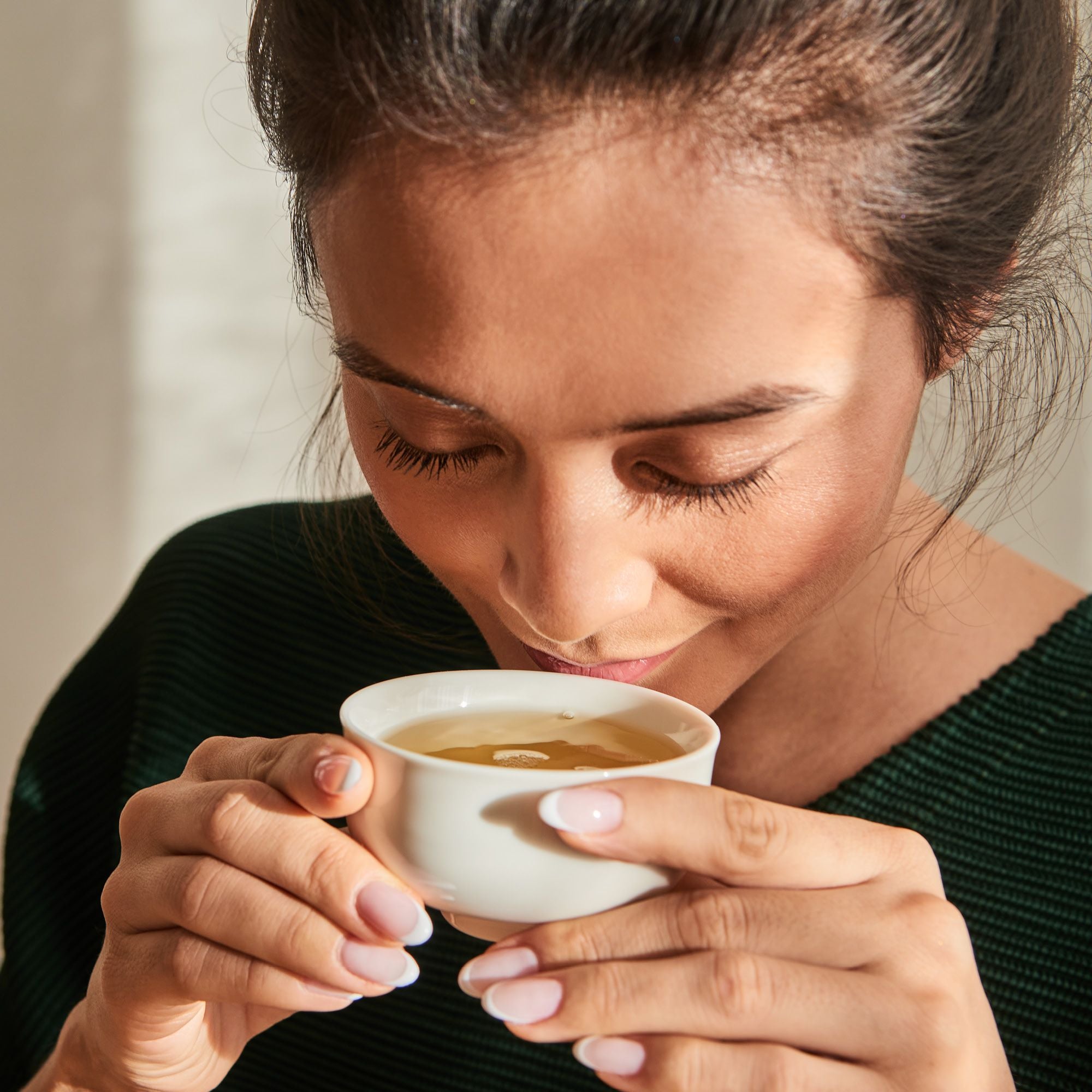 Frau in grünem Rippsweater trinkt entspannt Tee aus weißer Keramiktasse, zeigt French Nails.