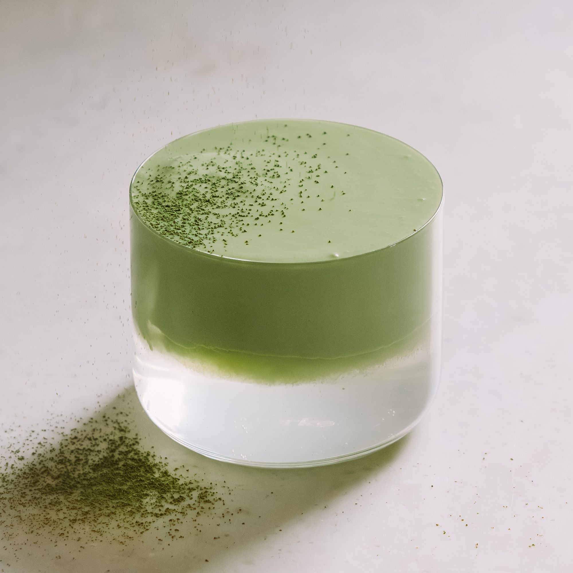 Ein zylindrisches Glas mit grüner und weißer cremiger Teeschichtung, bestreut mit grünem Teepulver.