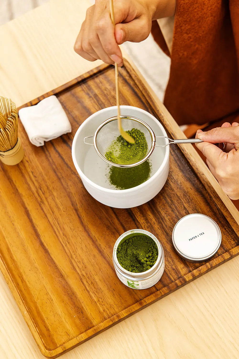 Hochwertiger Matcha-Tee mit Bambus-Sieb und Löffel auf Holztablett mit offener Matcha-Dose zubereitet.