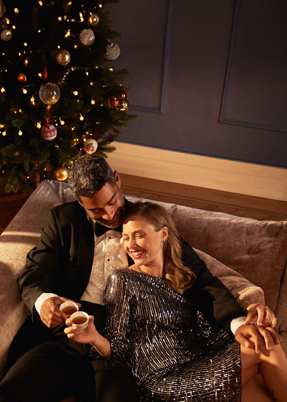 Romantisches Paar in eleganter Kleidung trinkt Tee vor festlich geschmücktem Weihnachtsbaum auf beigem Sofa.