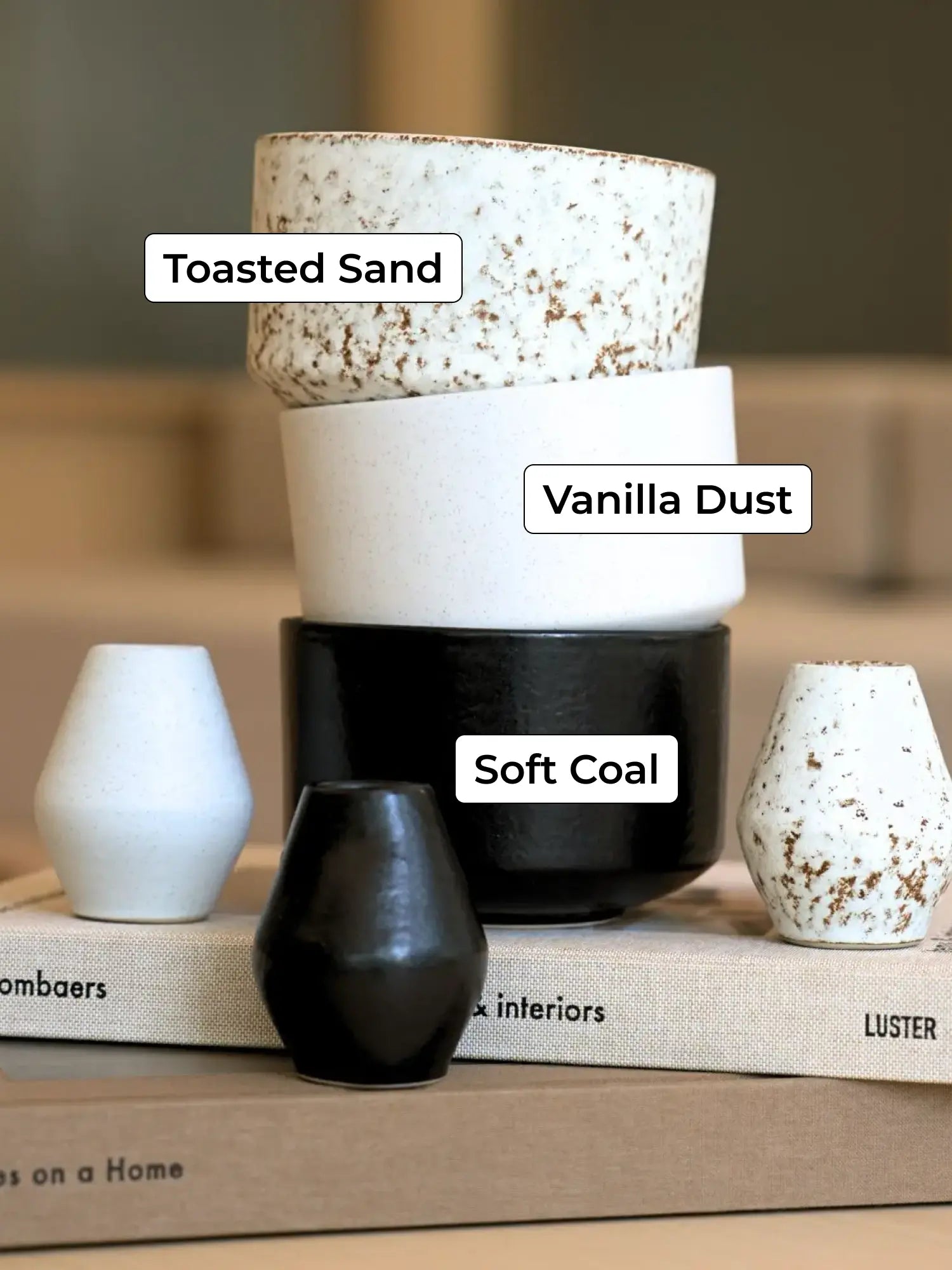 Drei keramische Schalen in Toasted Sand, Vanilla Dust und Soft Coal auf Büchern, umgeben von weißen Vasen.