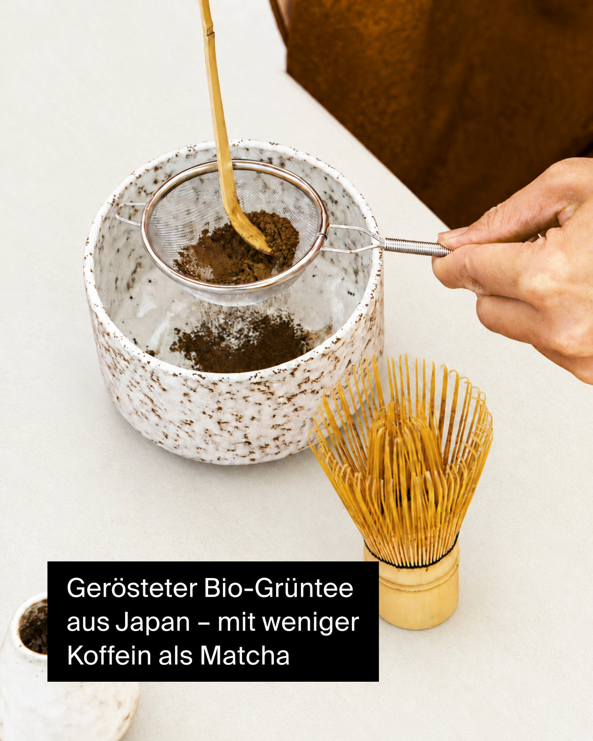 Hand hält Metallsieb mit geröstetem Bio-Grüntee über weißer Keramikschale, daneben Bambusbesen.