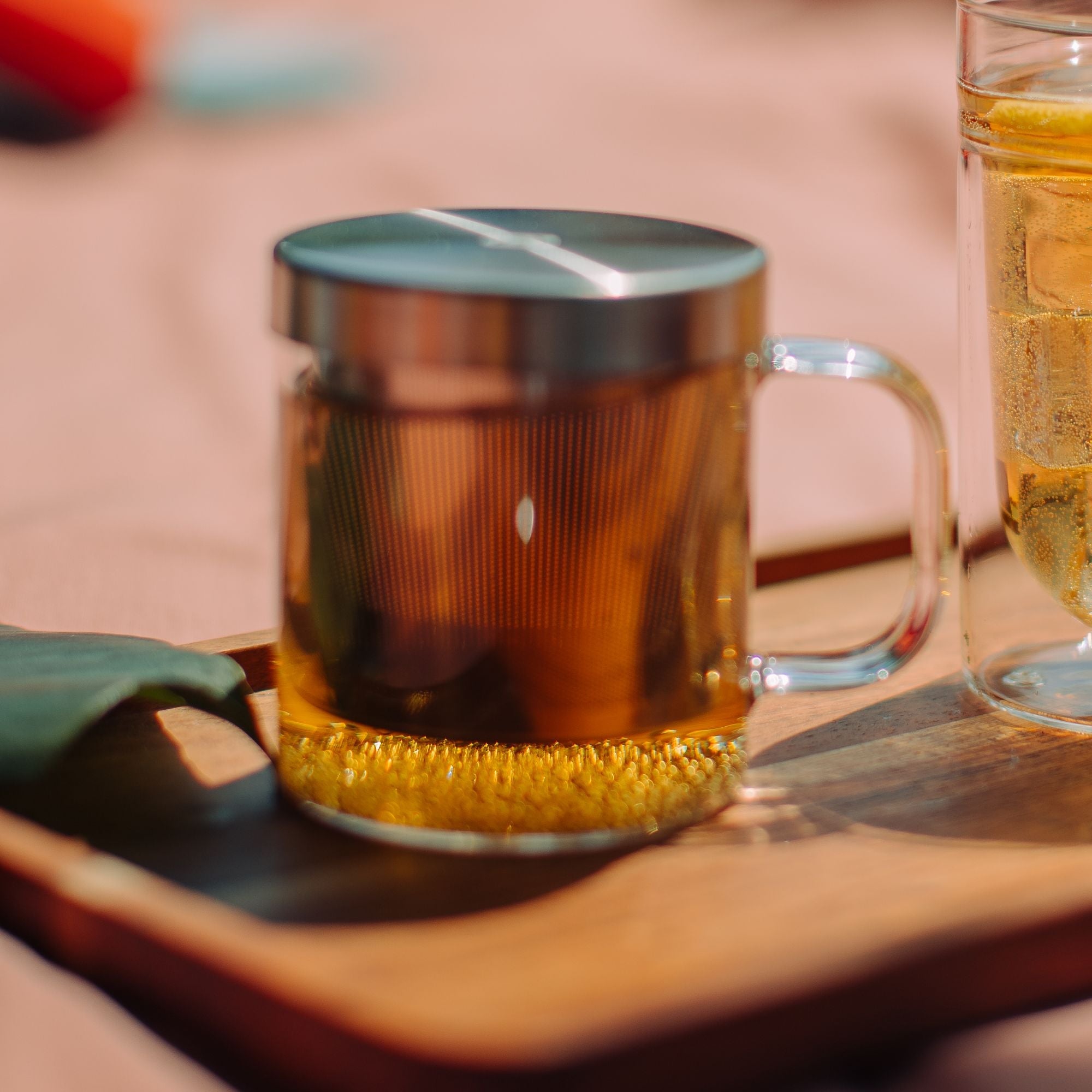 Glasbecher mit dunklem, bernsteinfarbenem Tee und Metalldeckel neben hellen Eistee mit Eiswürfeln und Zitrone auf Holztablett.