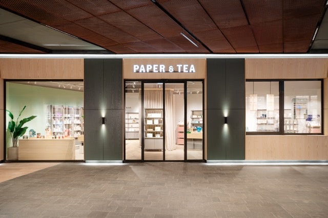 Moderner PAPER & TEA Laden mit Glasfronten, beleuchteten Regalen und grünen Pflanzen im Interieur.