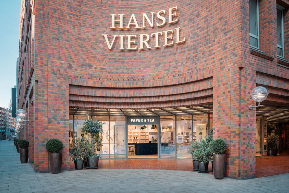 Eingang des P&T Ladens im Hanse Viertel mit goldenen Buchstaben und symmetrischen Pflanzen am Backstein.