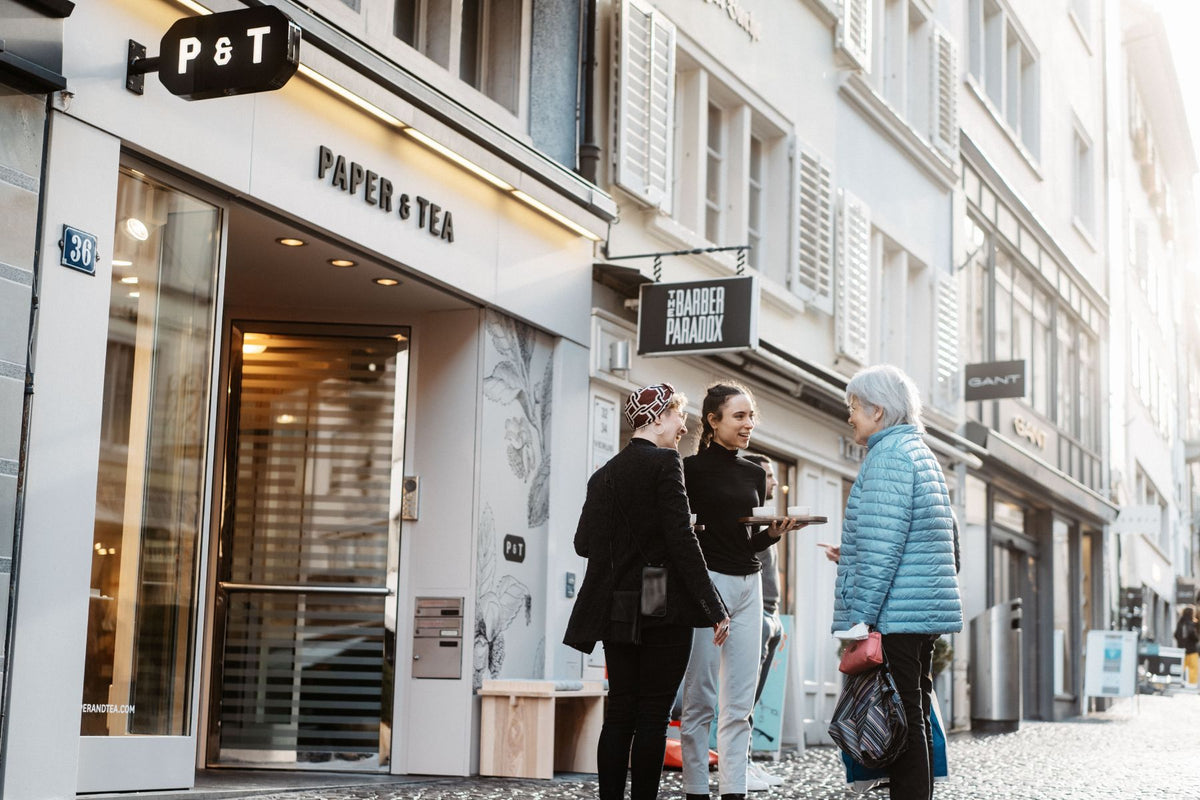 Straßenszene mit dem beleuchteten Eingang des P&T Teeladens Nr. 36, drei Menschen im Gespräch auf Kopfsteinpflaster.