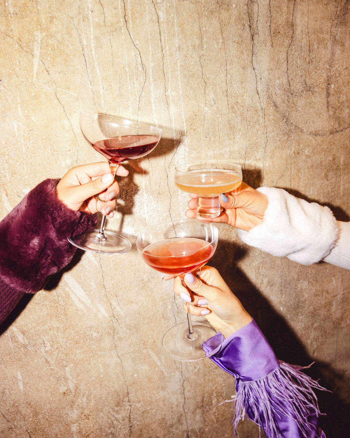 Drei Hände stoßen mit Cocktailgläsern in Rot, Gelb und Rosa vor beiger Wand an.
